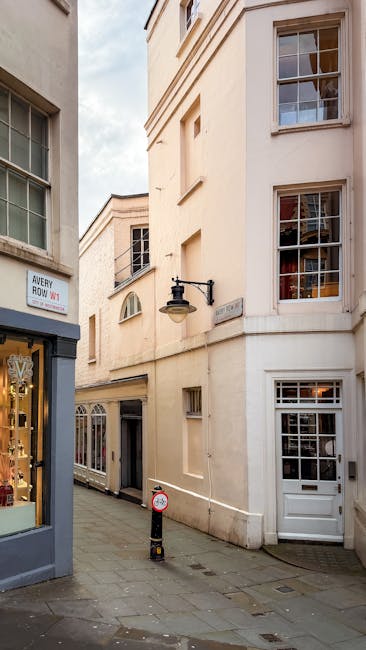 A narrow urban street scene in Mayfair showing cream-colored buildings with classic sash windows, some with decorative window panes, and a white door at street level. The pavement is made of cobblesto