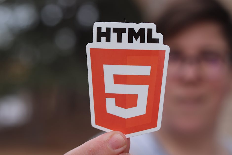 A person is holding a sticker featuring the HTML5 logo, which consists of a shield shape in orange and white with the text 'HTML5' at the top. The background appears to be a blurred garden scene with 