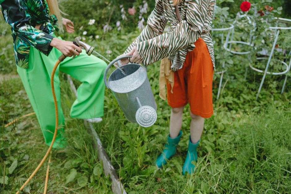 Two children are engaged in garden maintenance in a lush, green outdoor space. One child, dressed in bright green pants and a patterned jacket, is holding a garden hose connected to a spray nozzle, wa